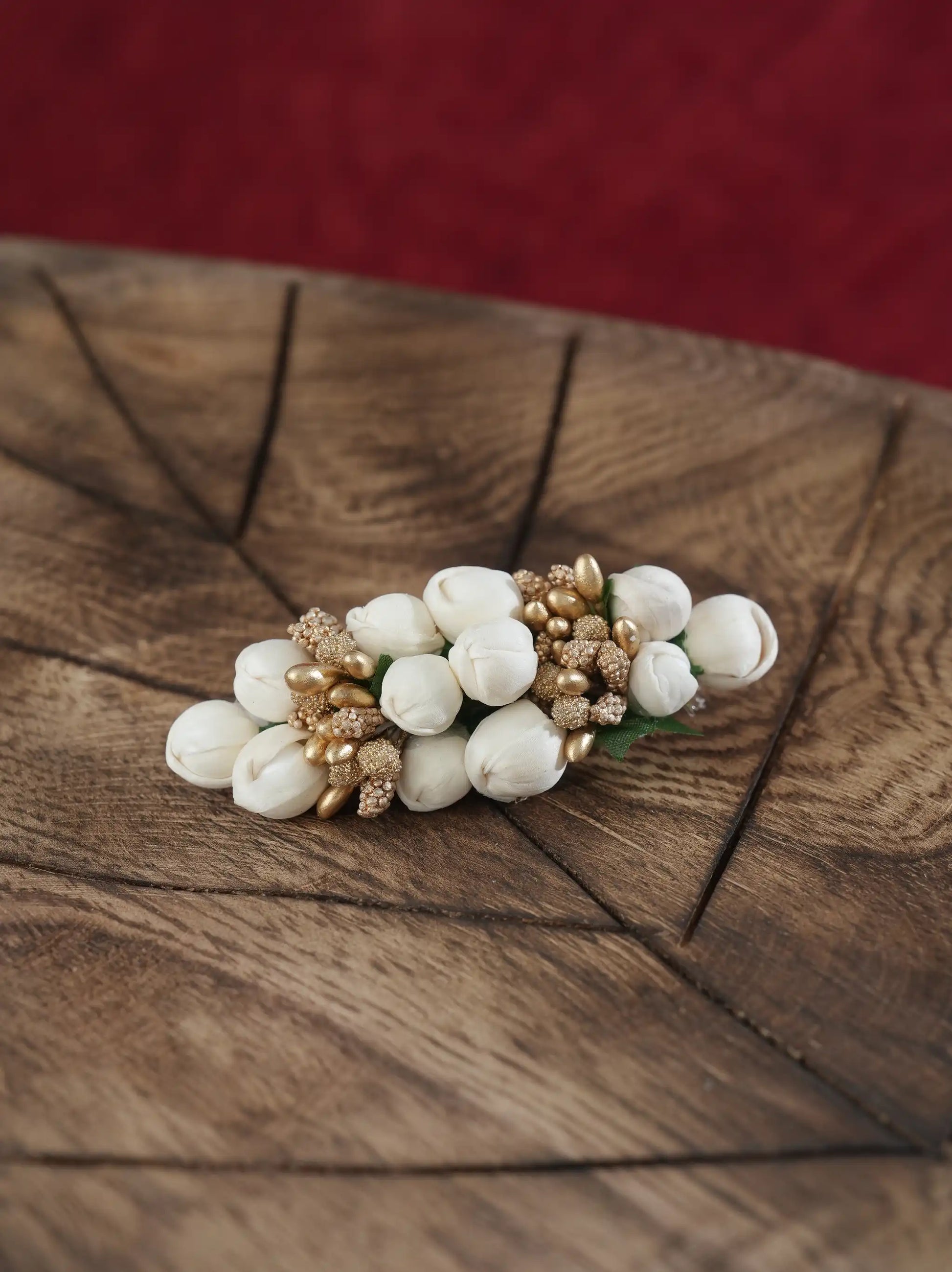 A small floral hair accessory with white tulip-like blossoms and metallic gold textured accents, displayed on a rustic wooden surface with a dark red background.