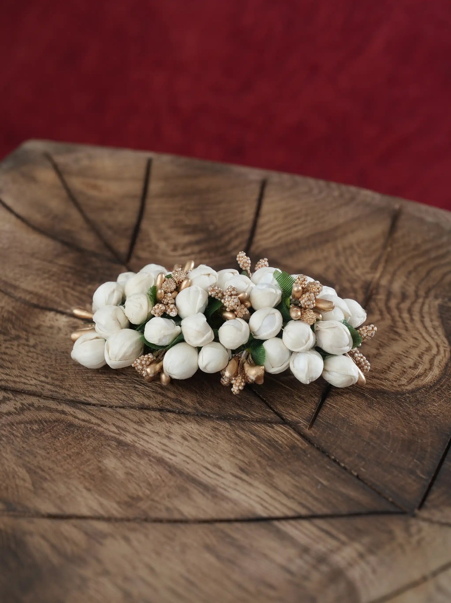 A white jasmine flower hair clip with gold-toned metal accents and small green leaves, displayed on a rustic wooden slice with a red textured background.