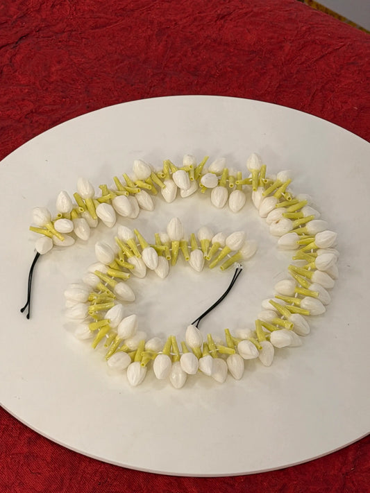 Two white flower lei necklaces with yellow-green stems and black elastic cords, arranged on a white oval plate over a textured red fabric background.