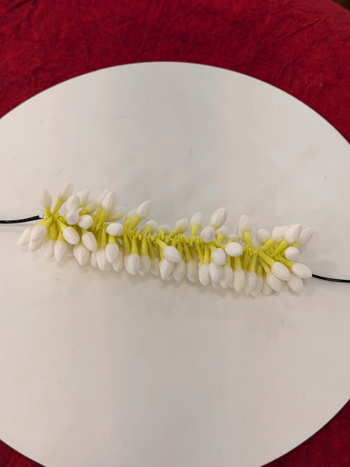 A white faux flower garland with yellow-green stems and black elastic bands, displayed horizontally on a round white plate against a red textured background.