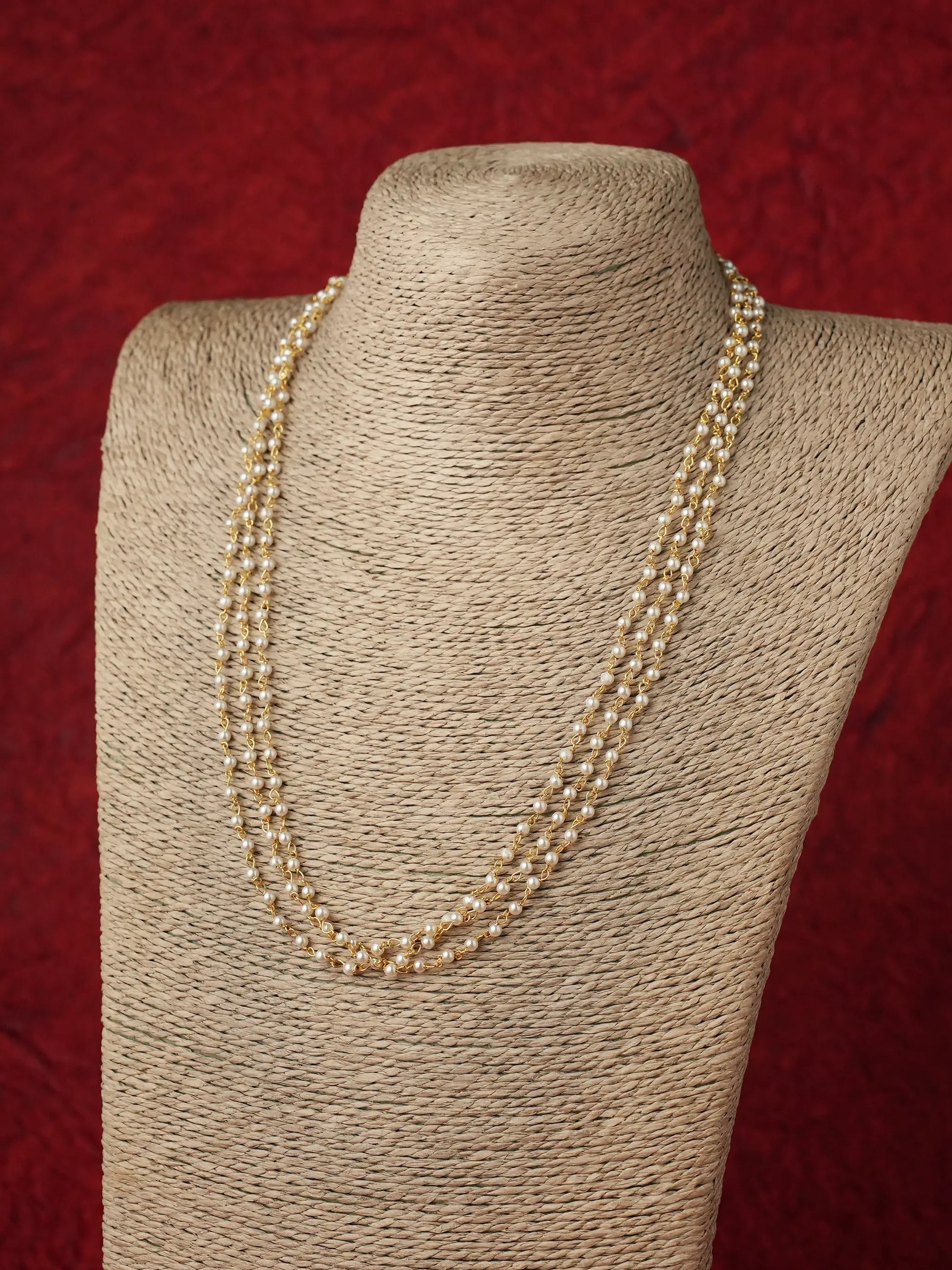 A multi-strand pearl necklace displayed on a beige fabric-covered jewelry bust against a textured red background.