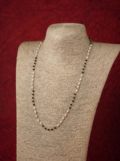 A long beaded necklace with small white pearls and dark beads, displayed on a textured beige fabric jewelry bust against a dark red background.