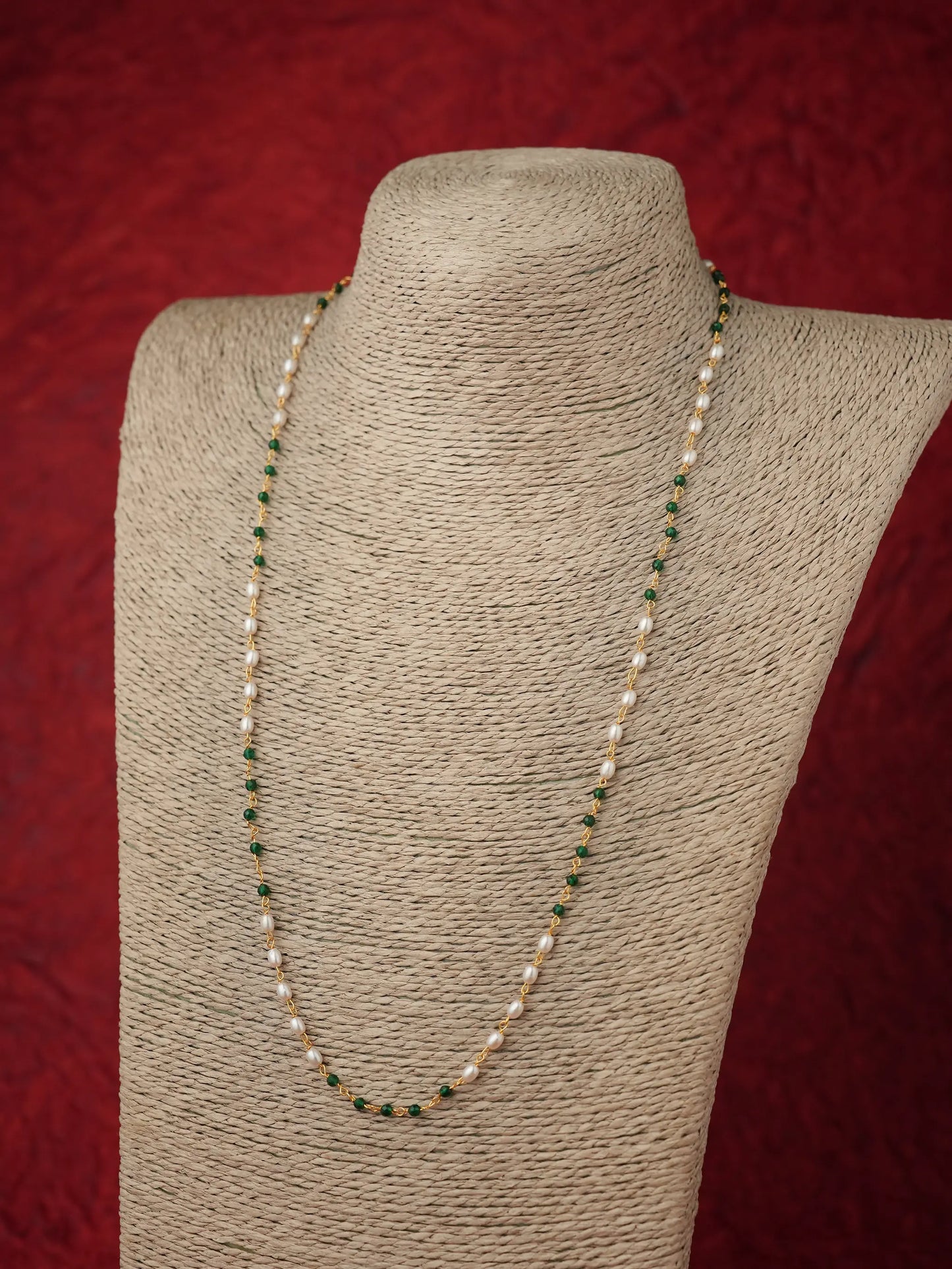 A beaded necklace with white pearls and small green beads, displayed on a beige fabric-covered jewelry bust against a textured red background.