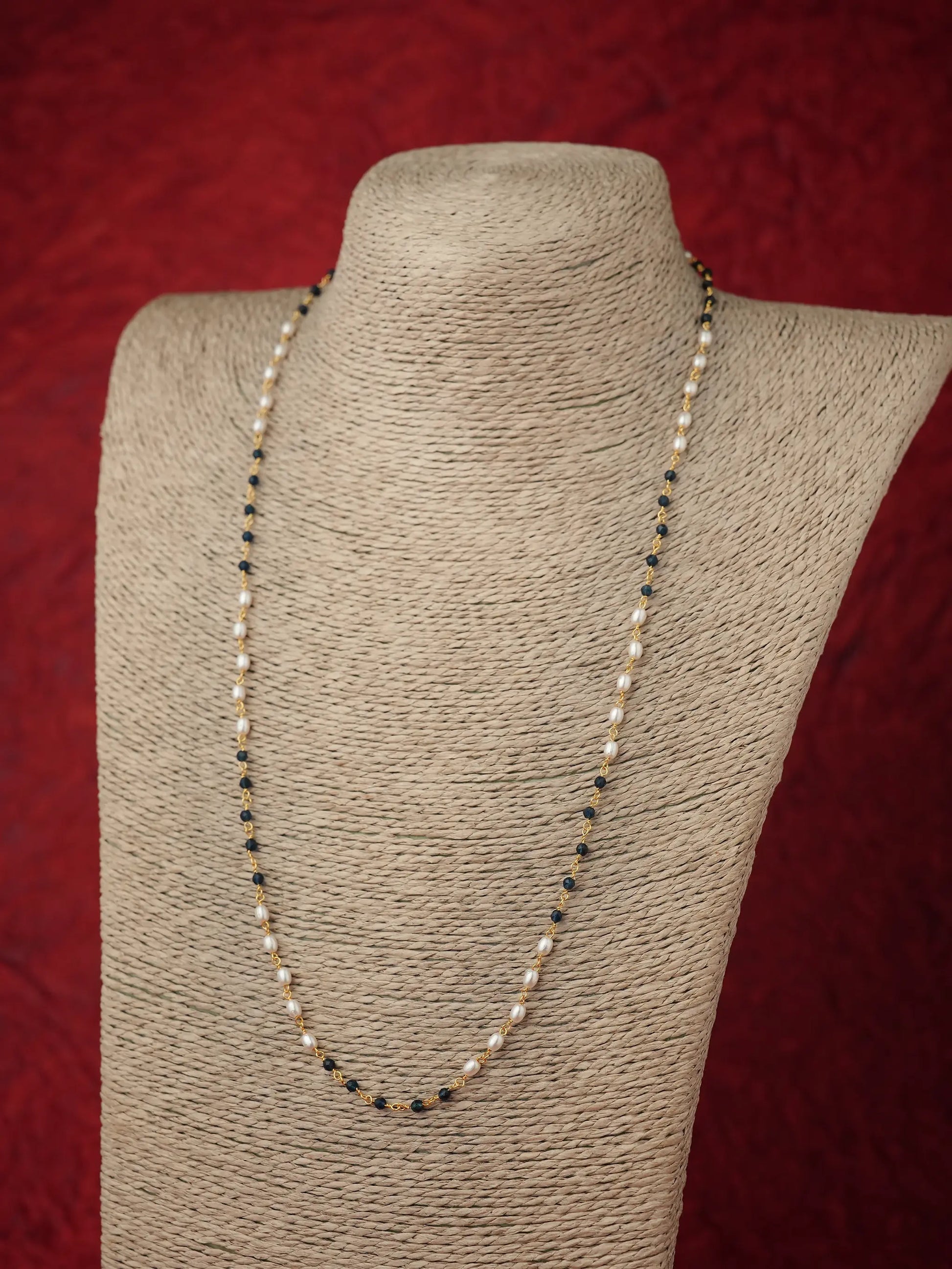 A long beaded necklace displayed on a textured beige fabric mannequin bust against a dark red background. The necklace features alternating strands of small white pearls and dark Blue beads separated by tiny metallic spacer beads.