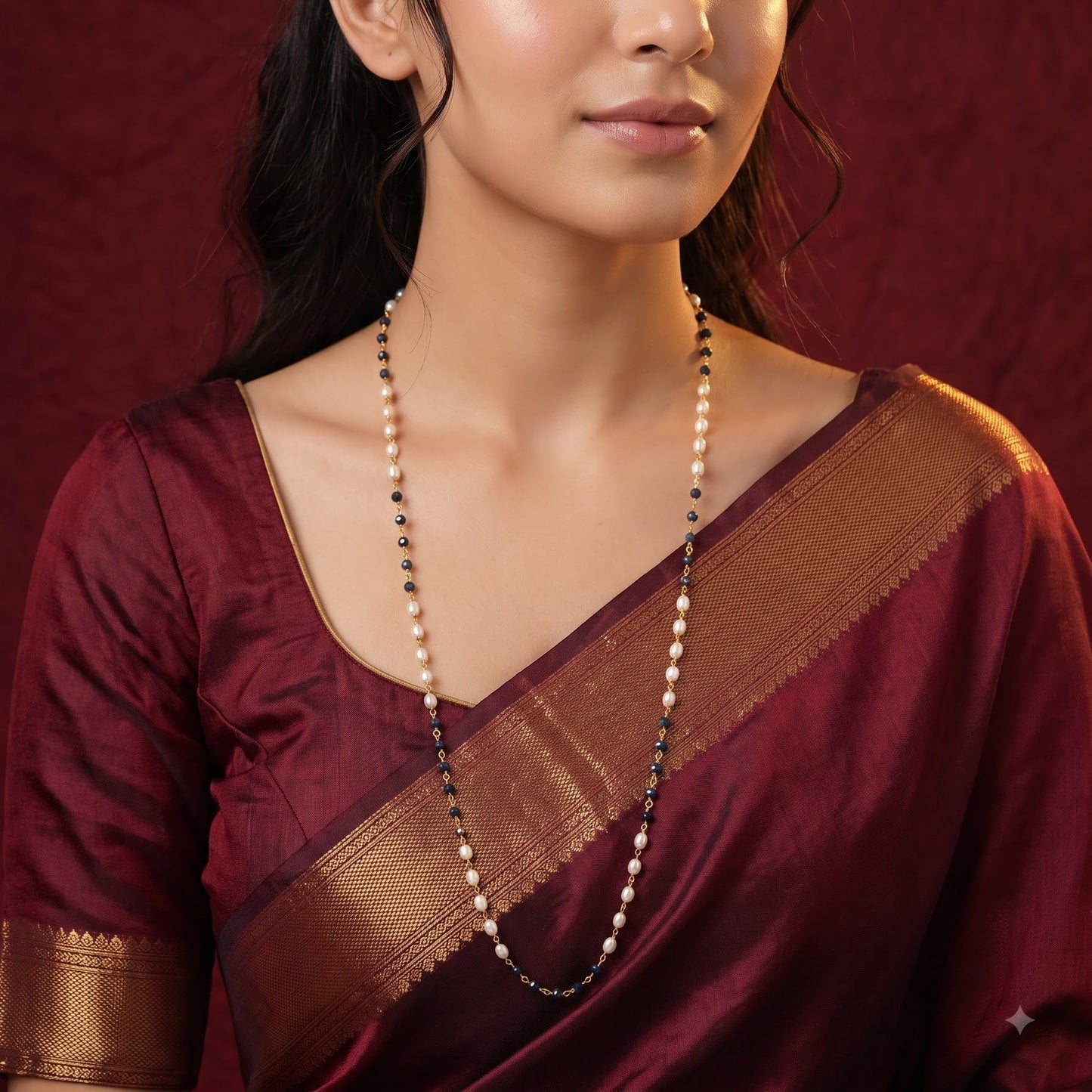 A woman wearing a deep maroon silk saree with a gold zari border and a matching maroon blouse, accessorized with a long beaded necklace, set against a dark red textured backdrop.