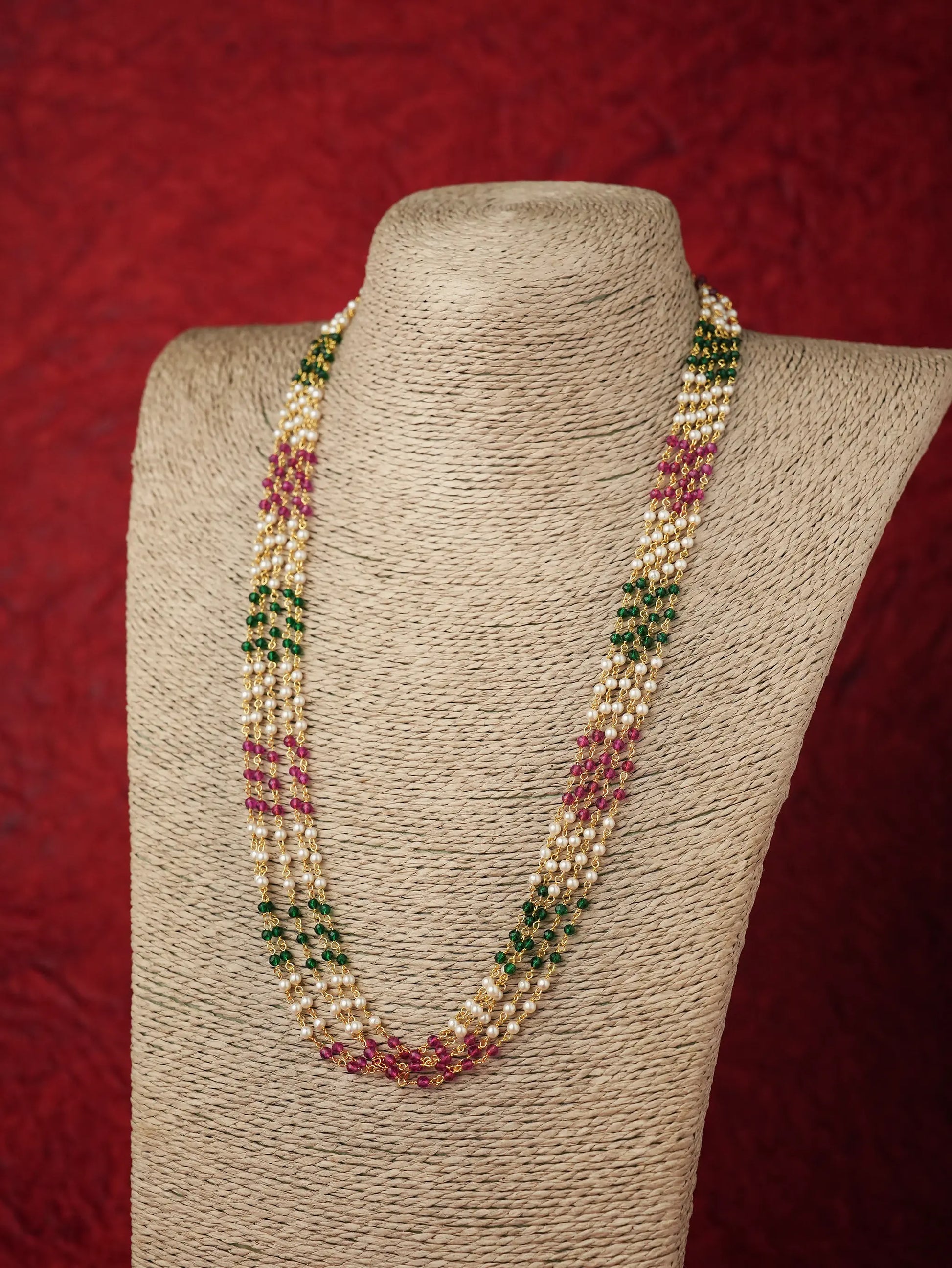 A multi-strand beaded necklace displayed on a textured beige fabric mannequin bust against a dark red background. The necklace features cream-colored beads, green beads, and pink beads woven together in a patterned design.