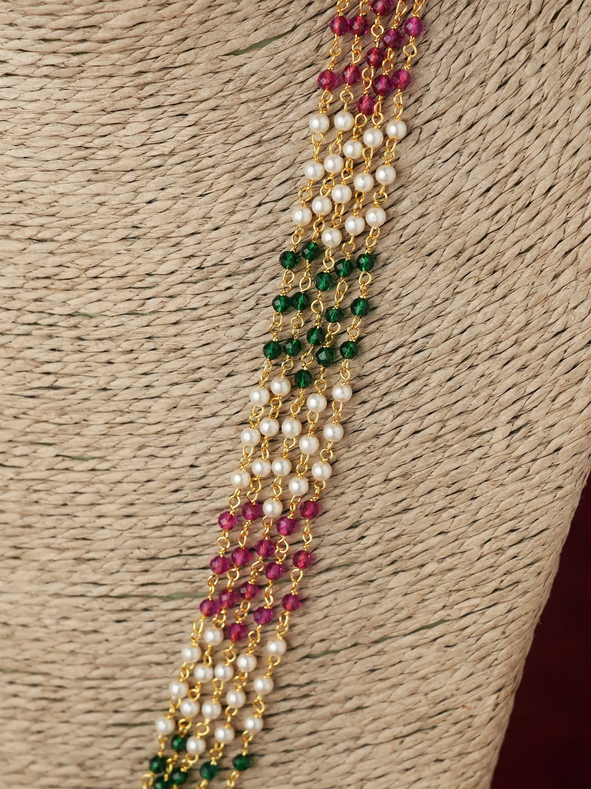A close-up of a multi-strand traditional Indian jewelry piece, likely a Kara or a necklace, featuring white pearls, green beads, and pinkish-red beads on a gold chain, displayed on a woven straw background.