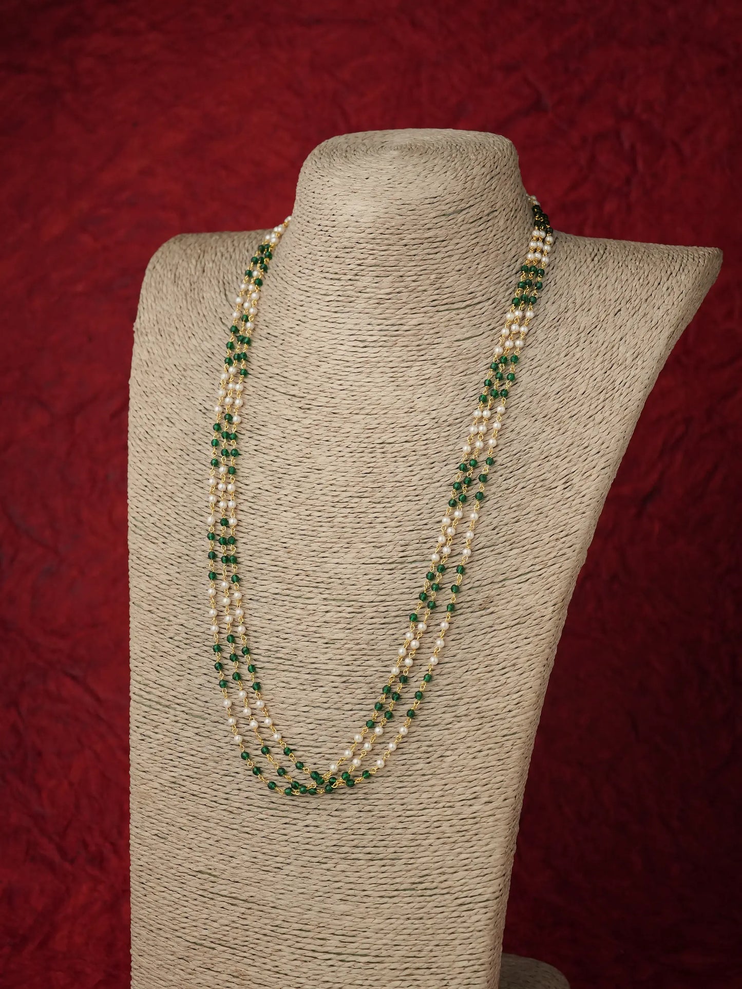 A multi-strand pearl necklace with green stone accents, displayed on a beige fabric-covered jewelry bust against a textured dark red background.
