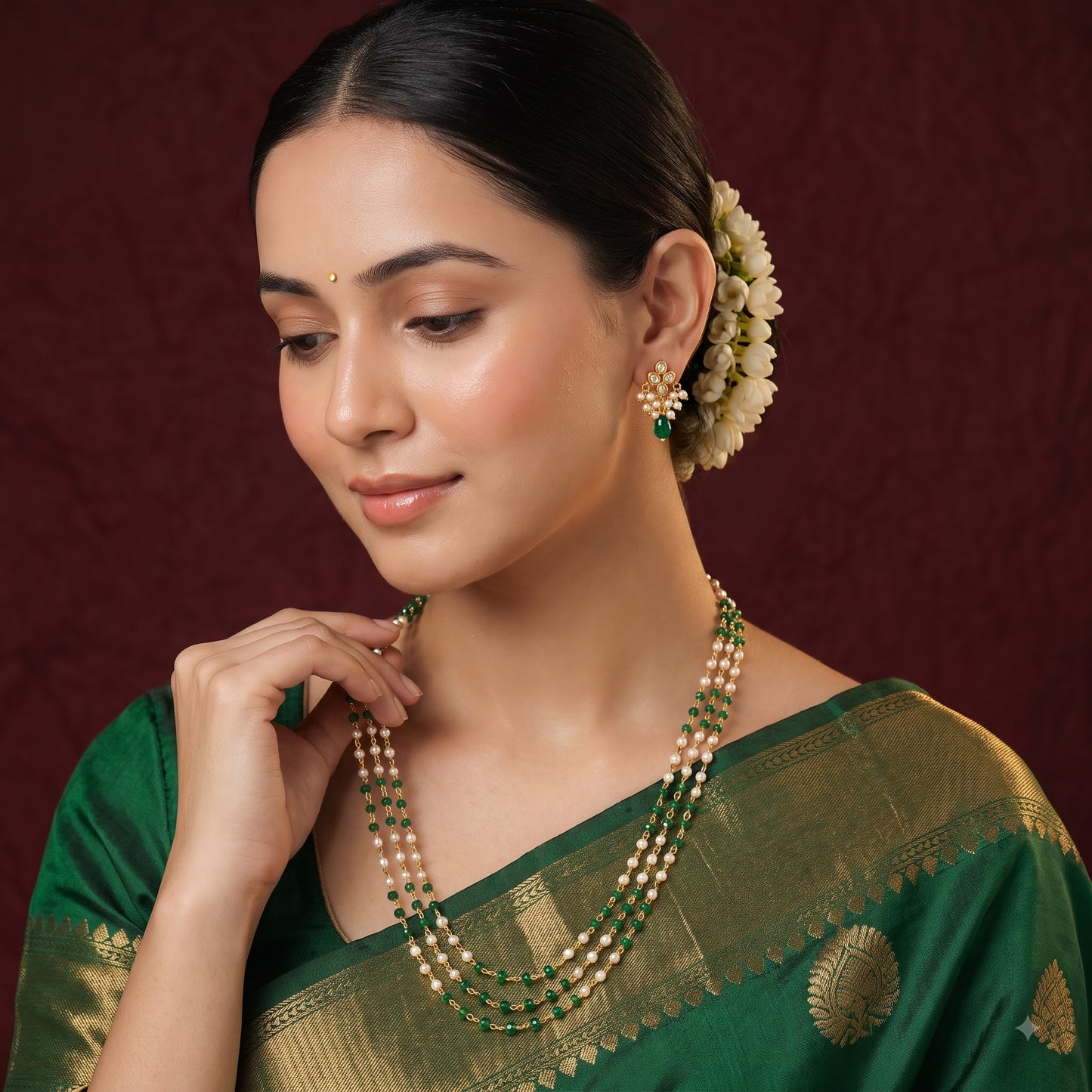 A woman wearing a green and gold sari with a matching beaded necklace and earrings, posed against a dark burgundy textured backdrop.