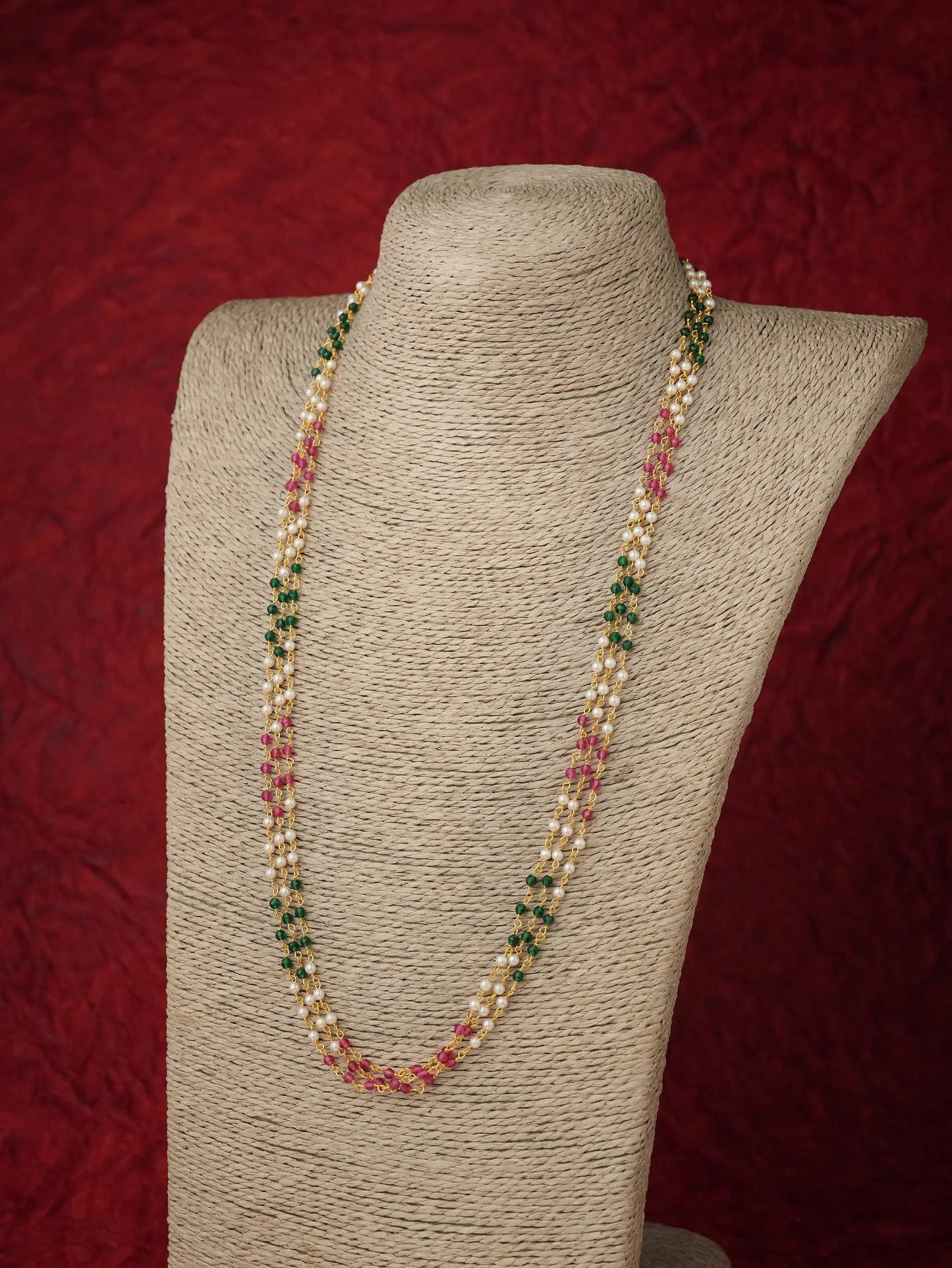 A long beaded necklace displayed on a textured beige fabric mannequin bust against a dark red background. The necklace features strands of white pearls interspersed with small green and pink beads.