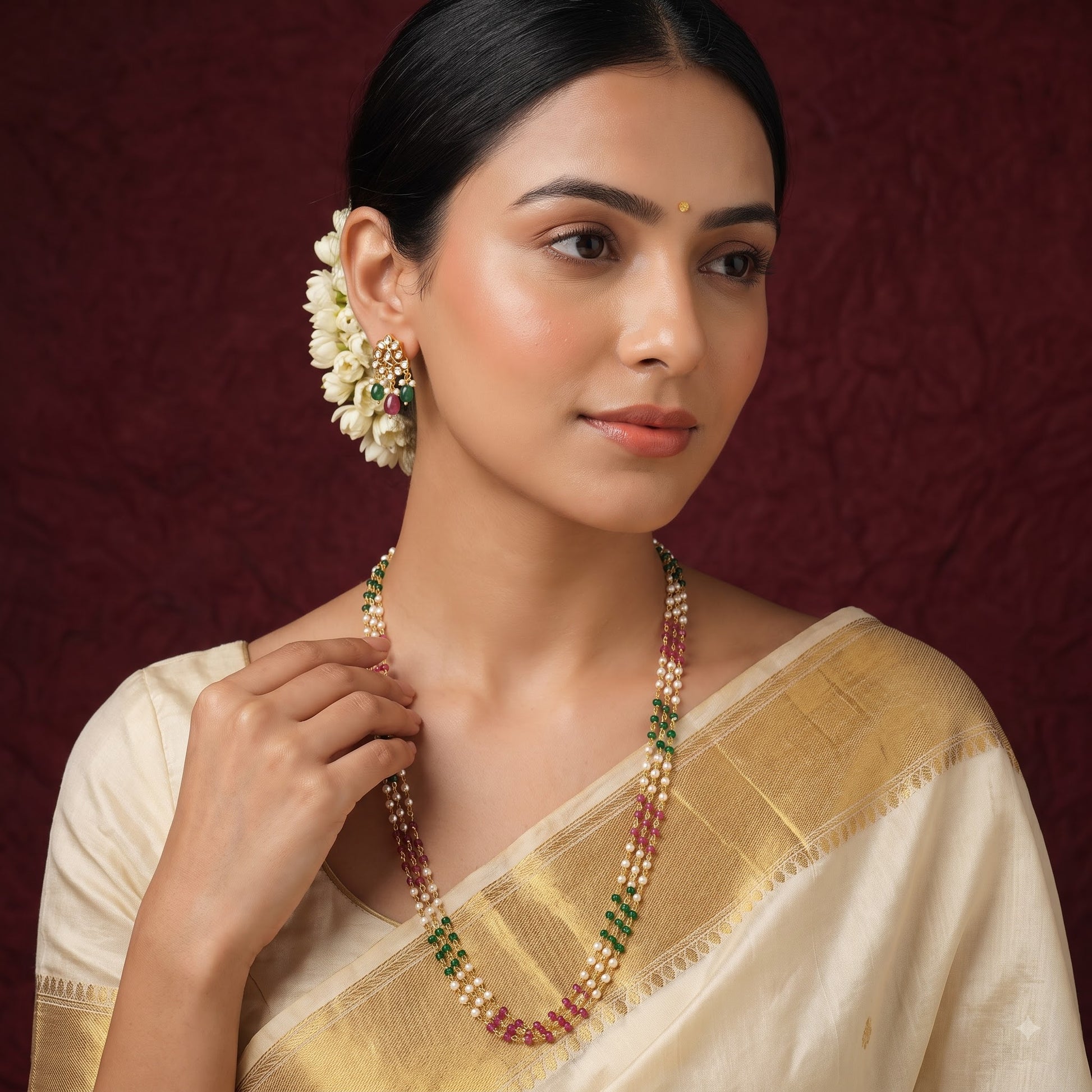 A woman wearing a traditional Indian outfit with a cream-colored saree and a gold border, accessorized with a beaded necklace and matching earrings. The jewelry features white pearls, red stones, and green stones, set against a dark burgundy textured background.