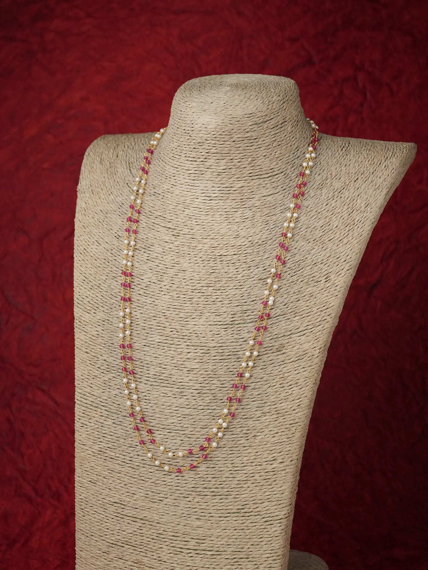 A long strand necklace with alternating cream pearls and small pink beads, displayed on a beige fabric-covered jewelry bust against a textured dark red background.