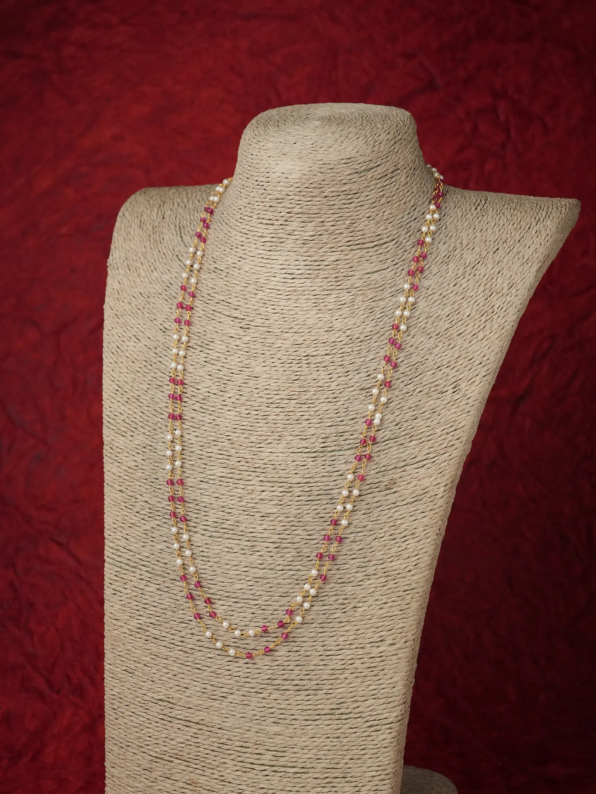 A long strand necklace with alternating cream pearls and small pink beads, displayed on a beige fabric-covered jewelry bust against a textured dark red background.
