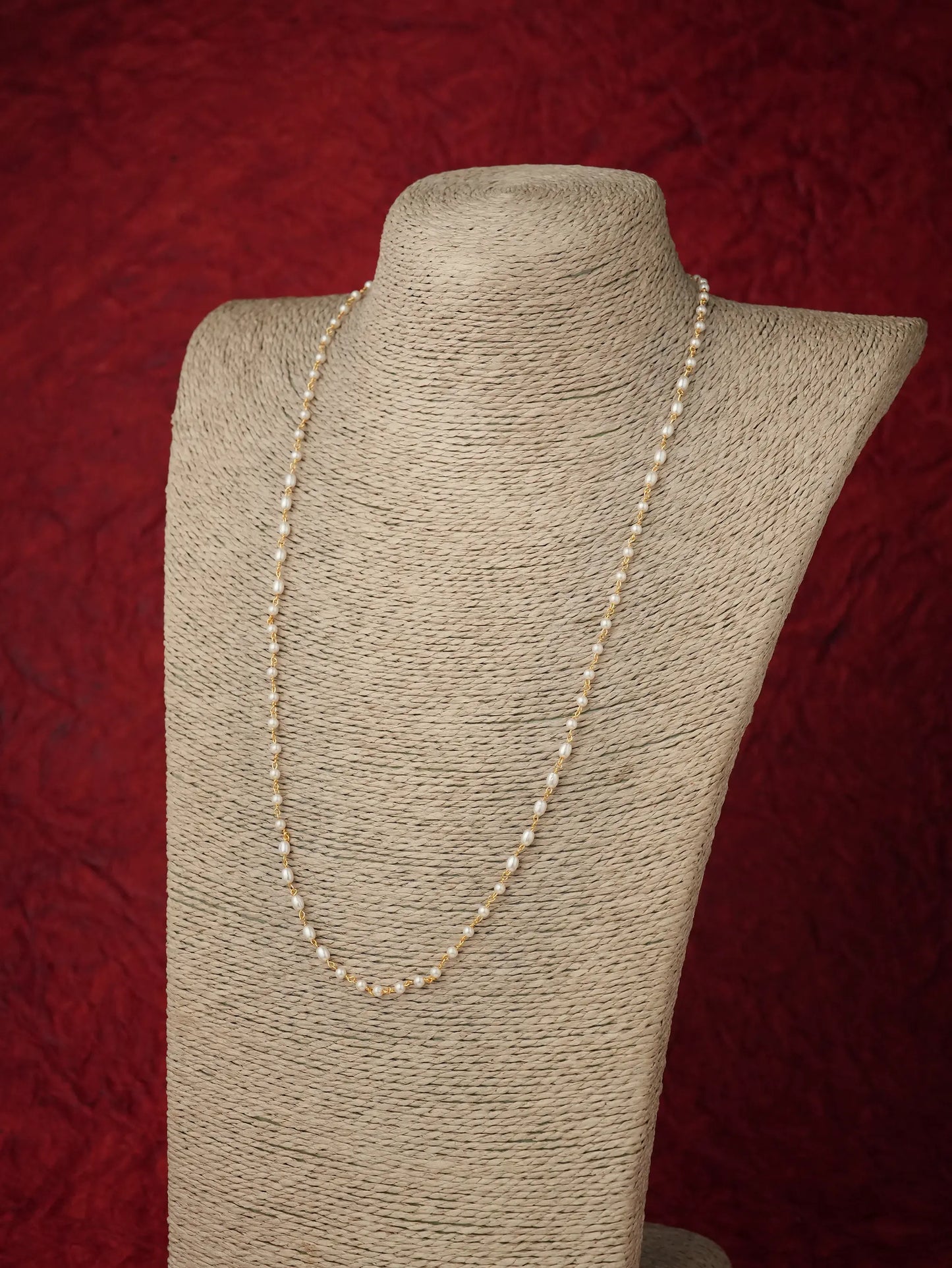 A long strand of cream-colored seed pearls displayed on a beige fabric-covered jewelry bust against a textured dark red background.