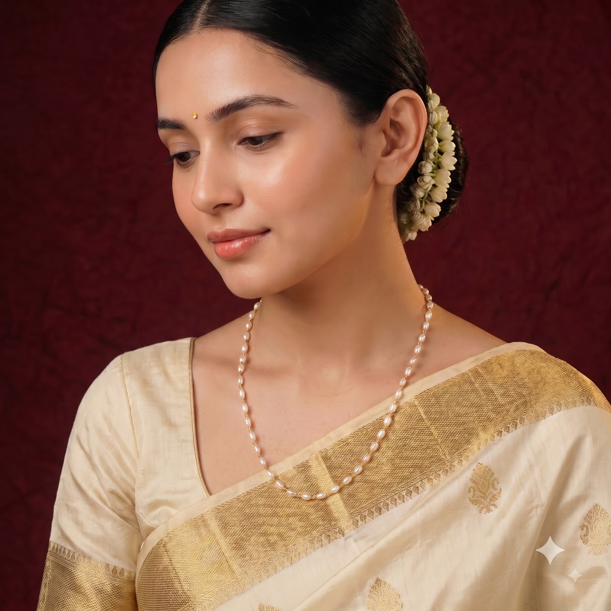 A woman wearing a cream-colored silk saree with a gold zari border and a matching pearl necklace, set against a dark red textured background.