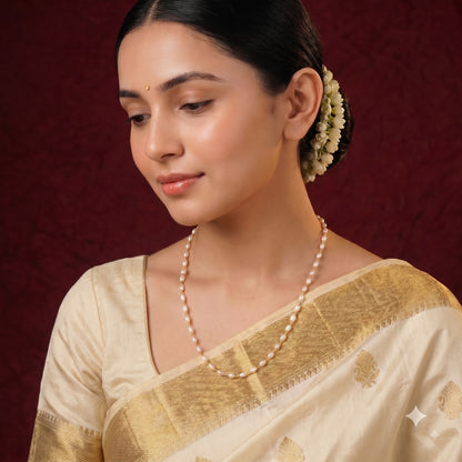 A woman wearing a cream-colored silk saree with a gold zari border and a matching pearl necklace, set against a dark red textured background.