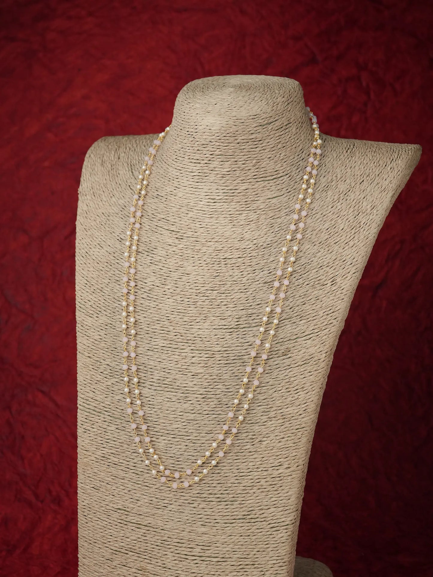 A long strand of cream and light pink colored pearls draped over a beige fabric-covered jewelry display bust against a textured dark red background.