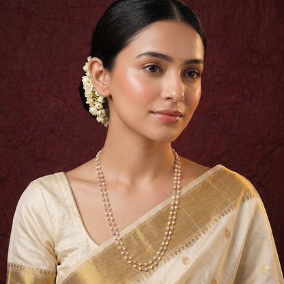 A woman wearing a cream and gold saree with a double-strand pearl necklace, set against a dark textured burgundy background.