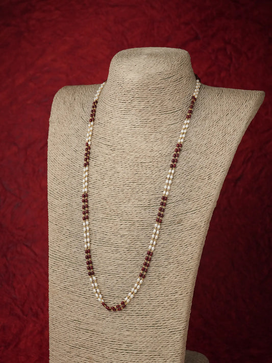 A long beaded necklace displayed on a textured beige fabric jewelry bust against a dark red textured background. The necklace features alternating strands of small white beads and dark reddish-brown beads.