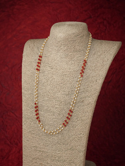 A long strand necklace with small cream pearls and alternating red faceted beads, displayed on a beige fabric-covered jewelry bust against a dark red textured background.