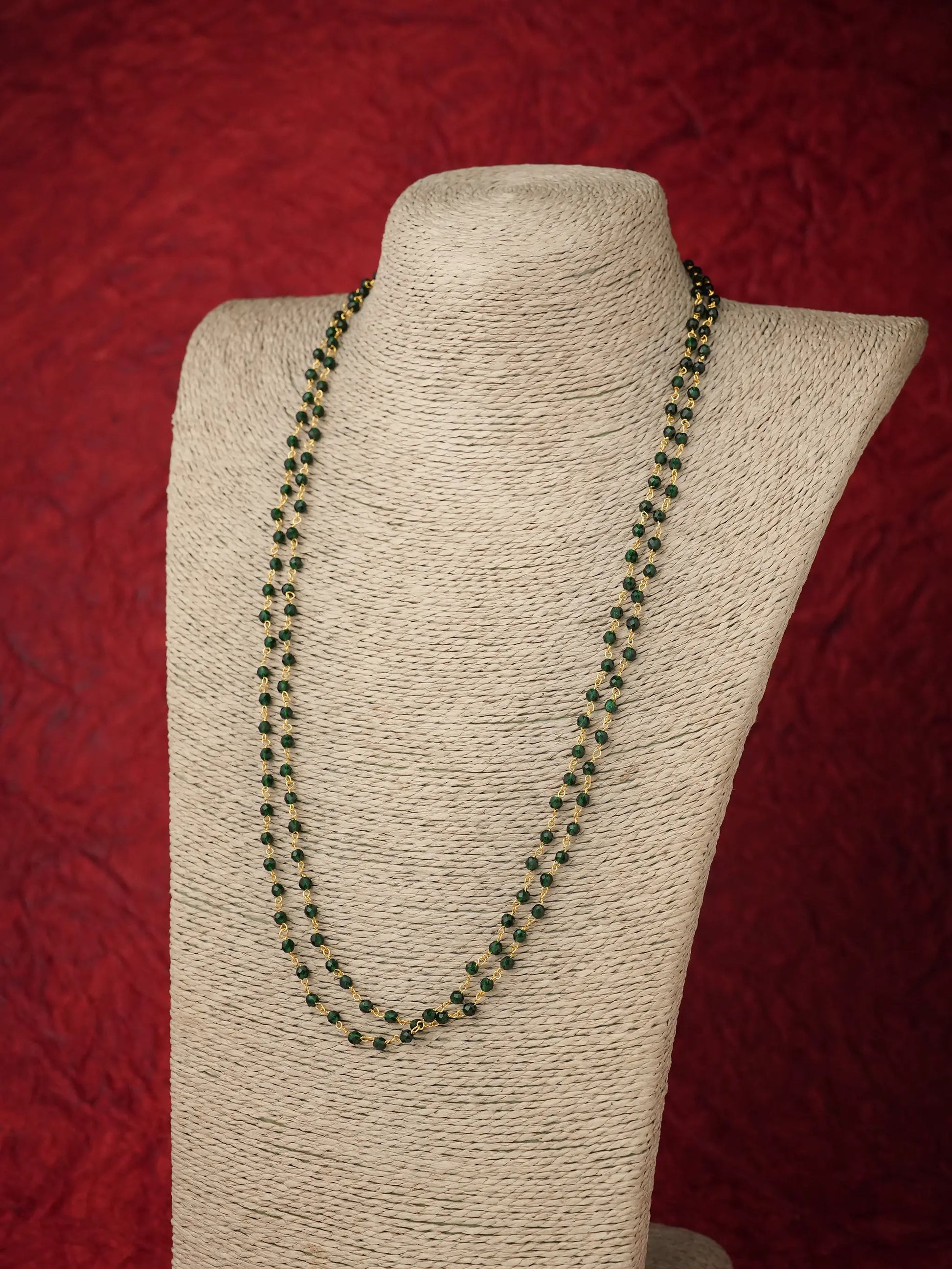 A double-strand beaded necklace with dark green and black beads, displayed on a beige fabric-covered jewelry bust against a textured red background.