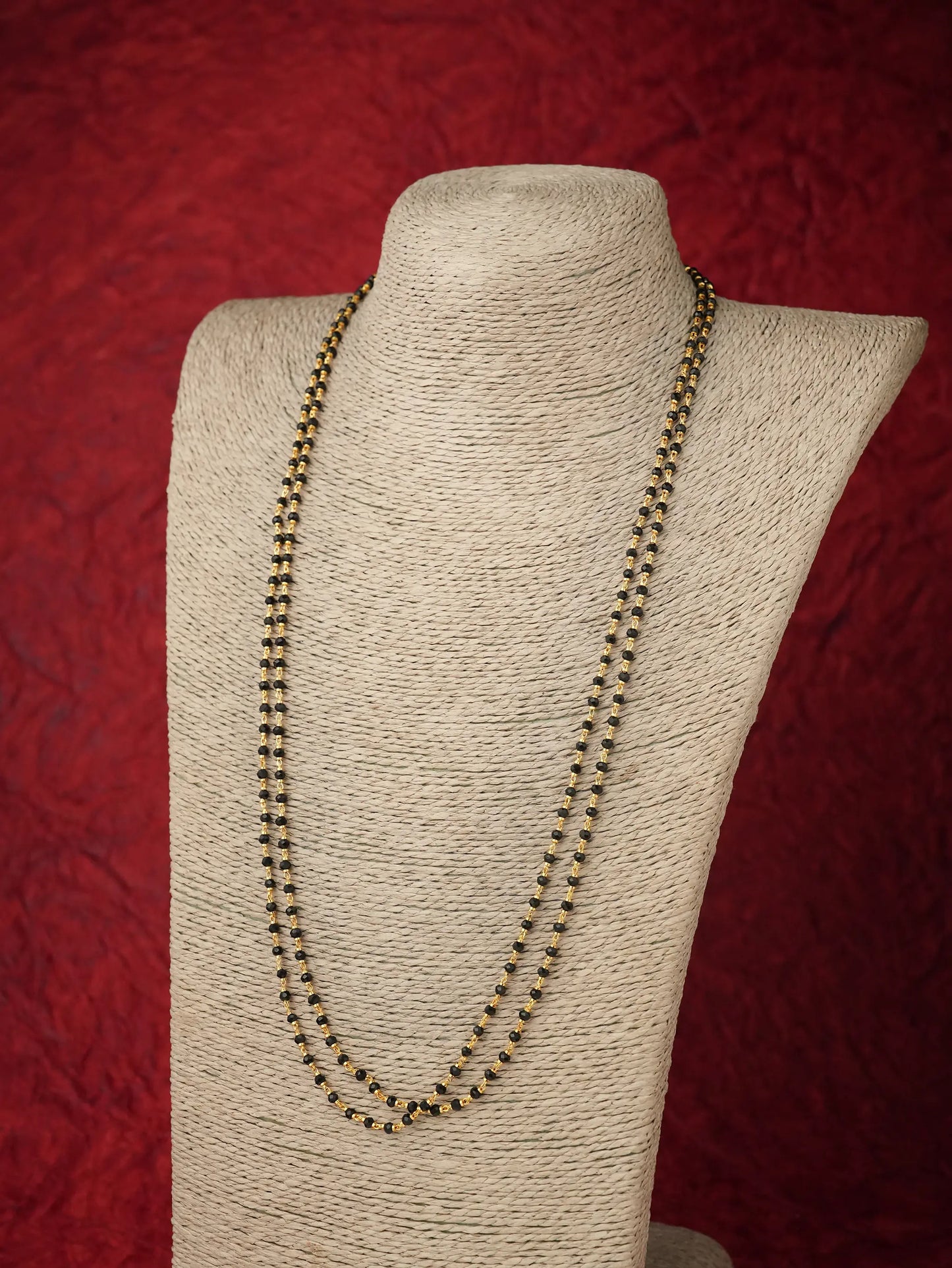A double-strand beaded necklace in a dark metallic bronze color, displayed on a textured beige fabric-covered jewelry bust against a textured red background.