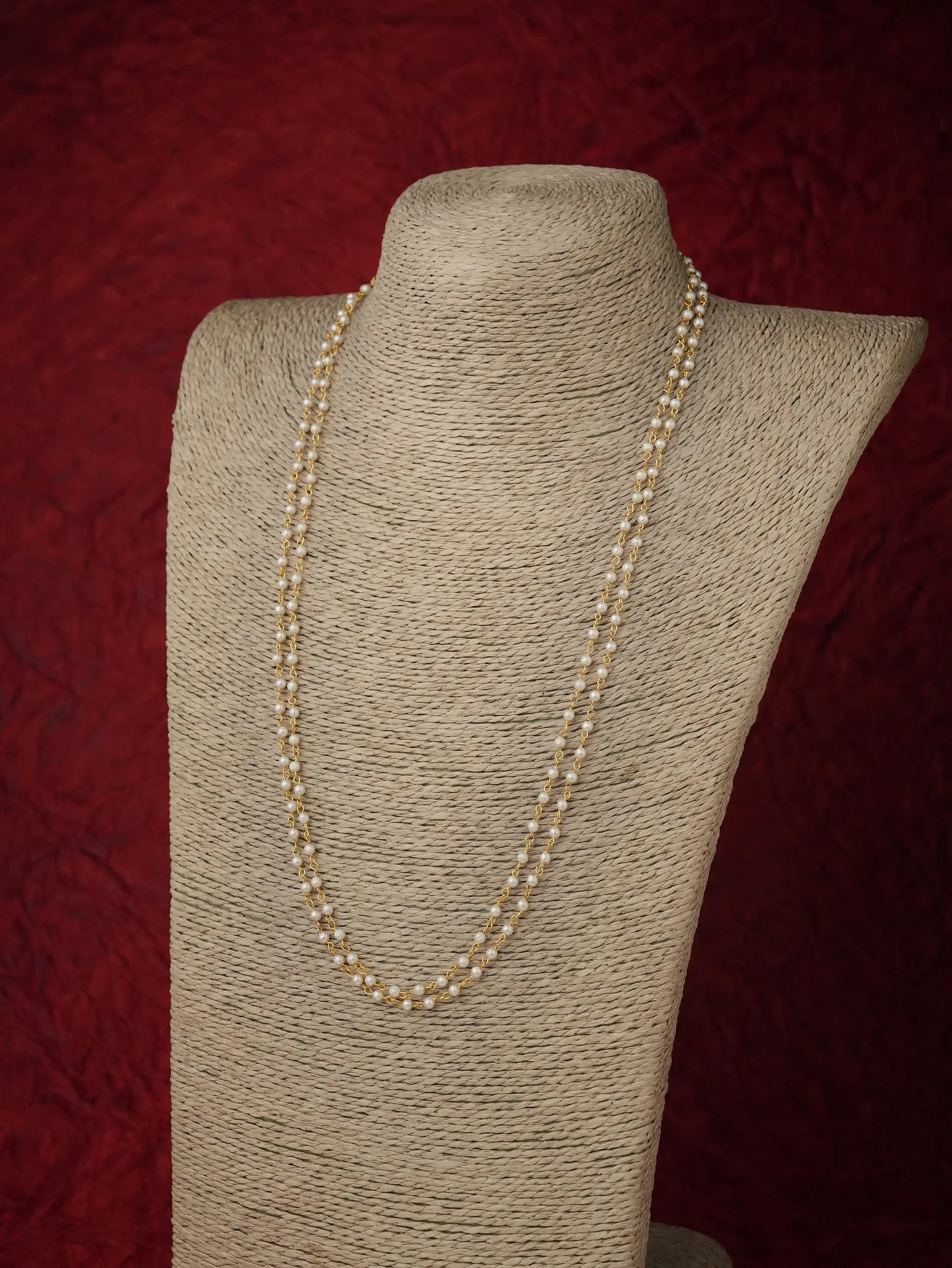 A long strand of cream-colored pearls draped over a beige fabric-covered jewelry display bust against a dark red textured background.