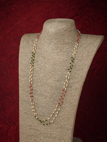 A double-strand pearl necklace with small green and red bead accents, displayed on a beige fabric-covered jewelry bust against a dark red textured background.