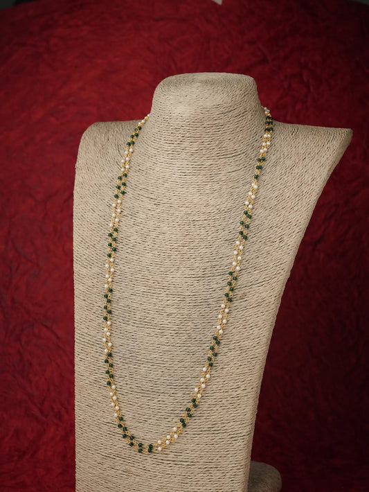 A long beaded necklace displayed on a beige fabric-covered jewelry bust against a dark red textured background. The necklace features alternating strands of small green beads and pale cream beads with gold-toned links.