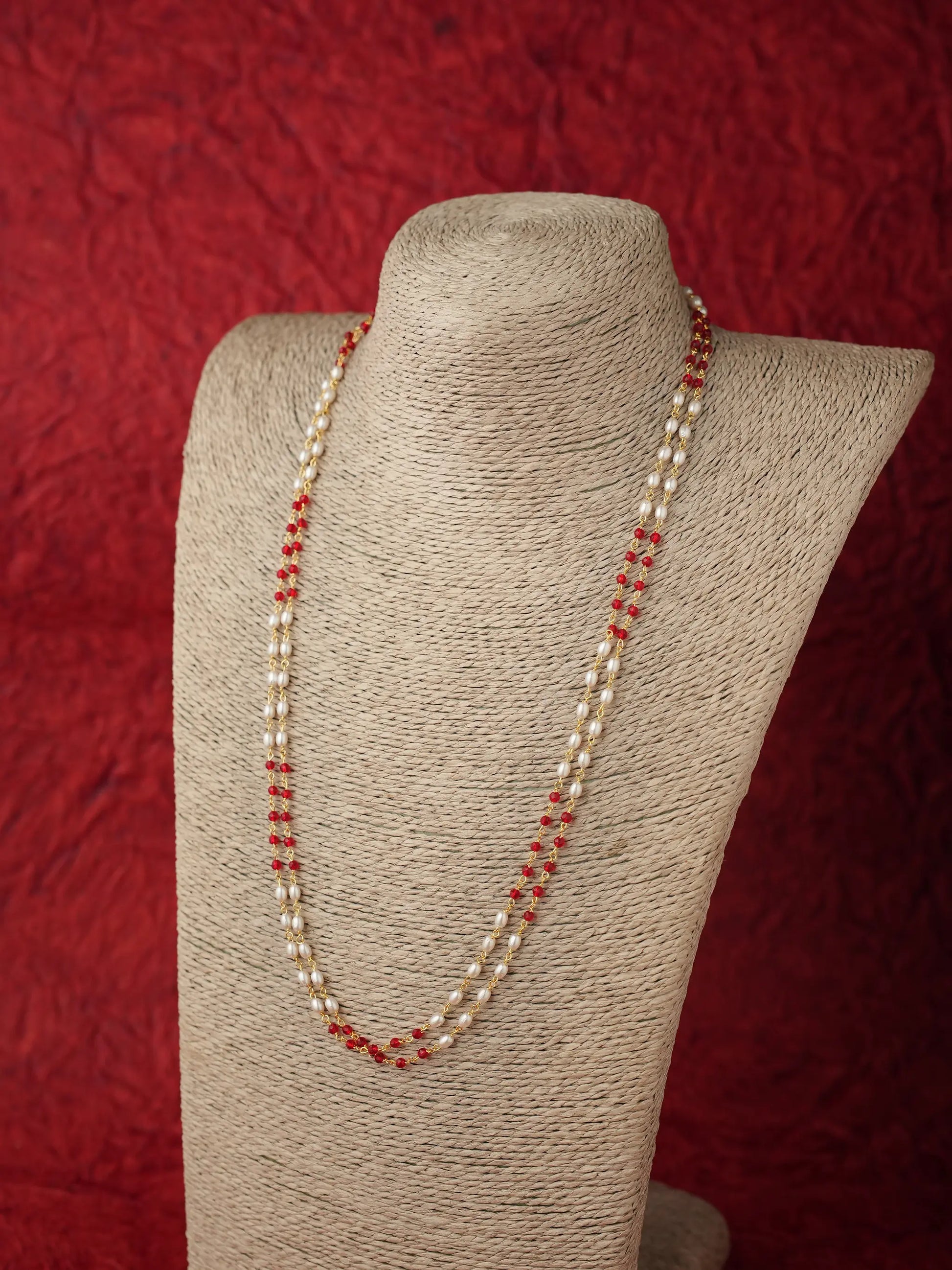 A double-strand necklace featuring white pearls and small red beads, displayed on a beige fabric-covered jewelry bust against a textured red background.