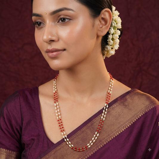 A woman wearing a traditional Indian outfit with a purple saree and a double-strand pearl and red bead necklace, posed against a textured dark red background.