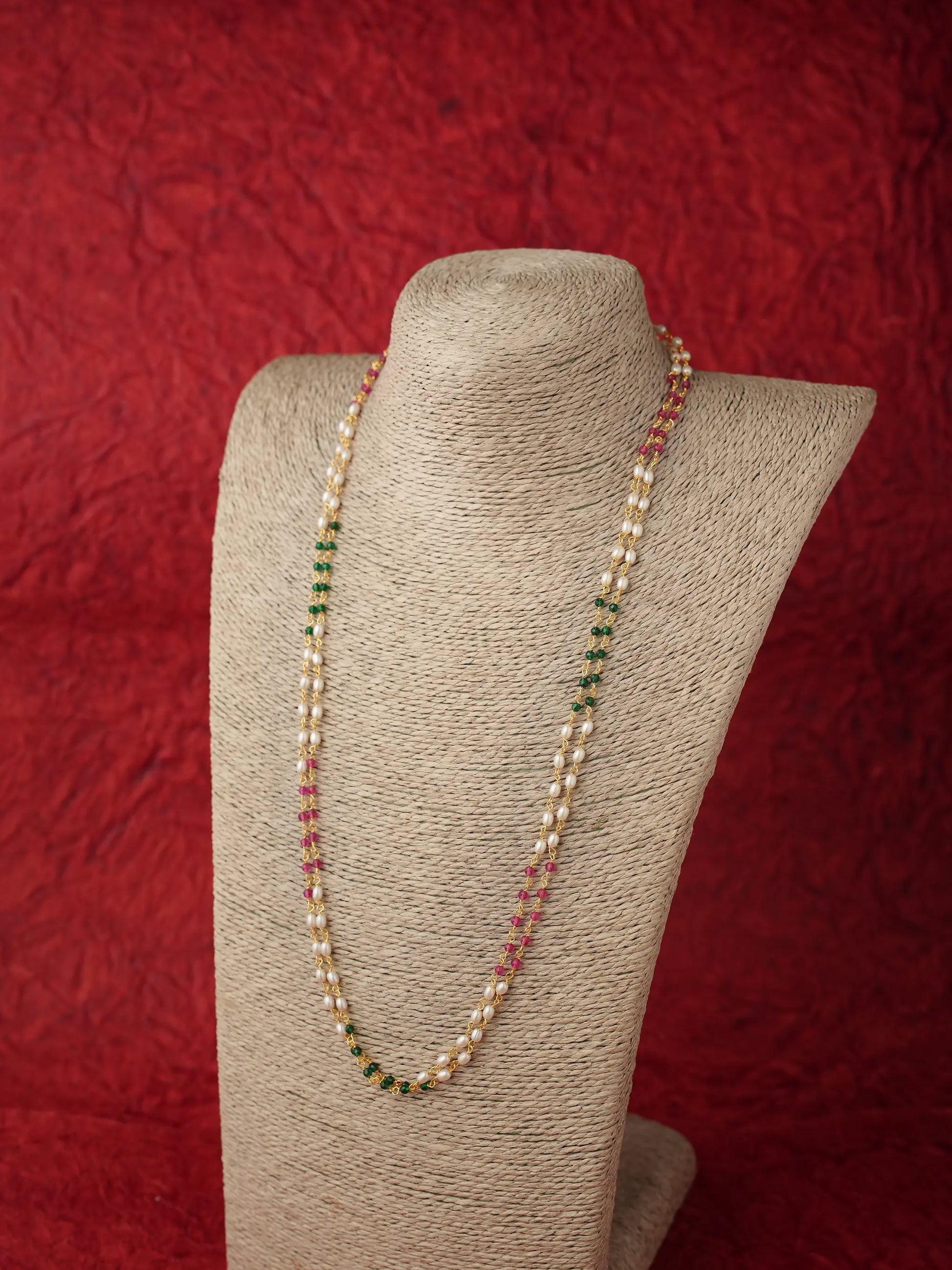 A long beaded necklace displayed on a beige fabric-covered jewelry bust against a textured red background. The necklace features alternating strands of small white pearls and green, pink, and red faceted beads.