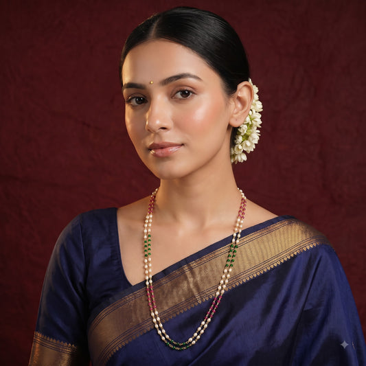 A woman wearing a navy blue silk saree with a gold zari border, styled with a beaded necklace and white flower hair accessory, set against a dark red textured background.