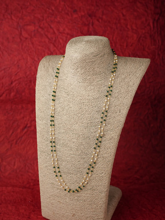 A long beaded necklace displayed on a textured beige jewelry bust against a red fabric backdrop. The necklace features cream-colored beads accented with small green beads.