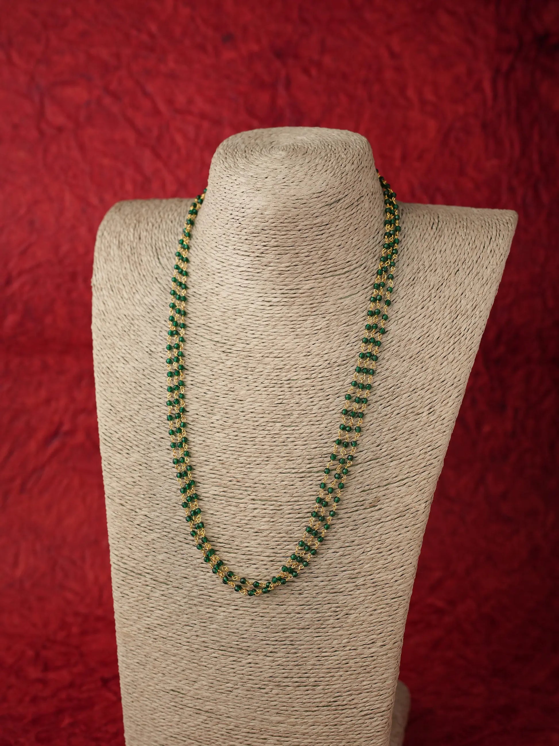 A green beaded necklace displayed on a beige fabric-covered jewelry bust against a textured red background.