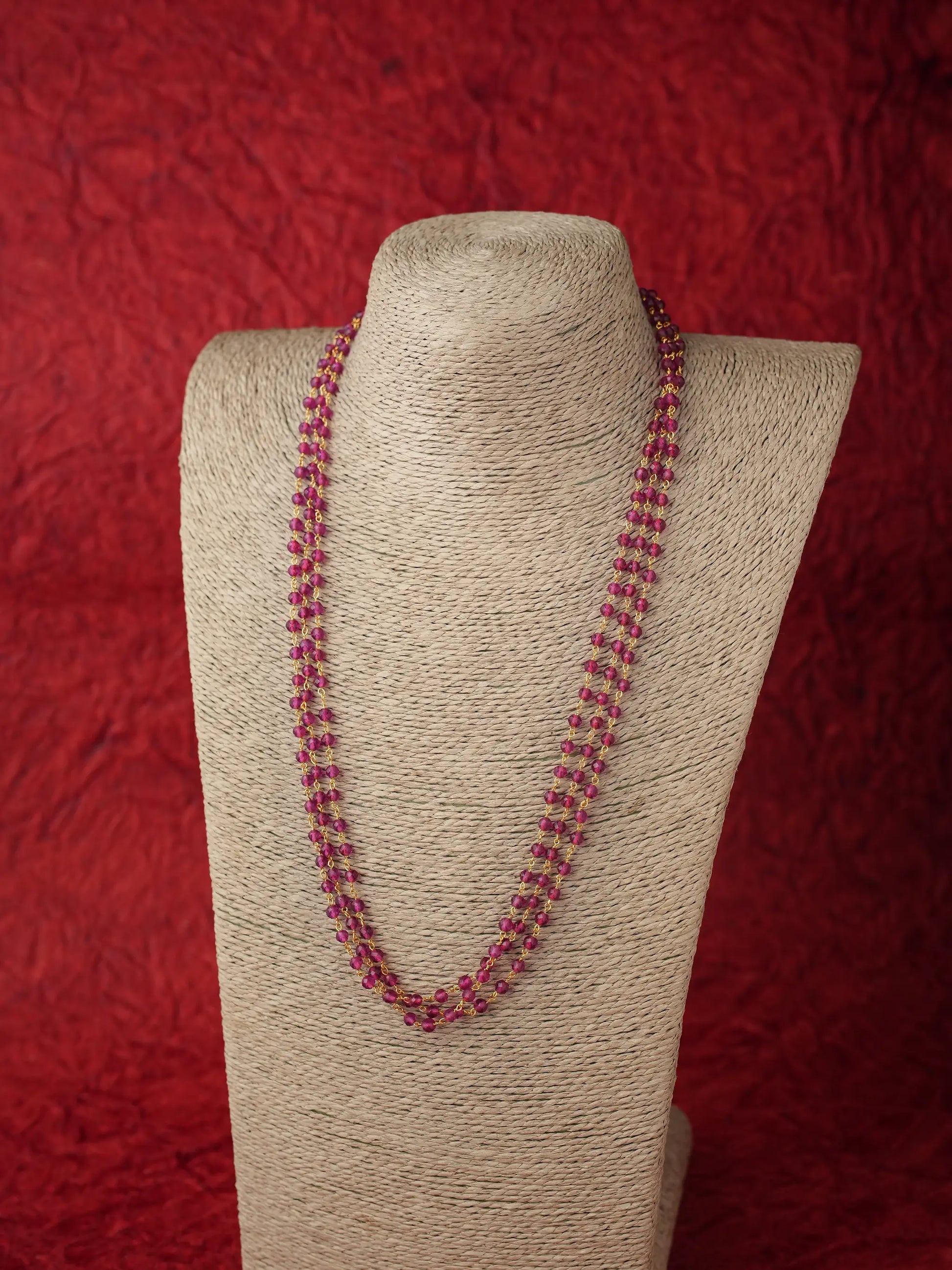 A beaded necklace with alternating red and clear beads, displayed on a beige fabric-covered jewelry bust against a textured red background.